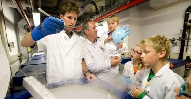 Drie kinderen en twee onderzoekers staan in een laboratorium te kijken naar een buisje dat één van de onderzoekers boven een bak met stoom houdt.