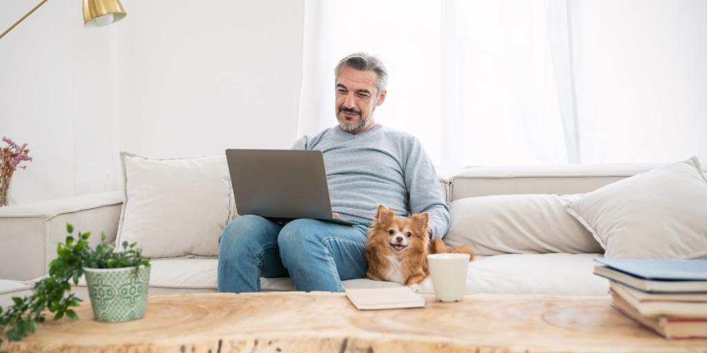 Man zit op bank met laptop, er ligt een hond naast de man