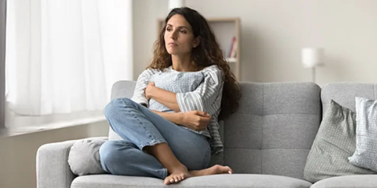 Een vrouw zit met haar benen opgetrokken op een grijze bank. Ze kijkt verdrietig.