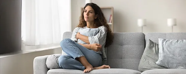 Een vrouw zit met haar benen opgetrokken op een grijze bank. Ze kijkt verdrietig.