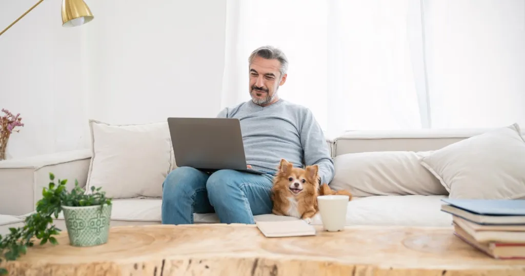 Man zit op bank met laptop, er ligt een hond naast de man