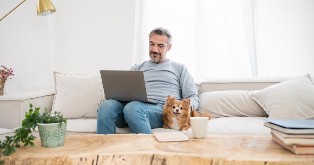 Man zit op bank met laptop, er ligt een hond naast de man